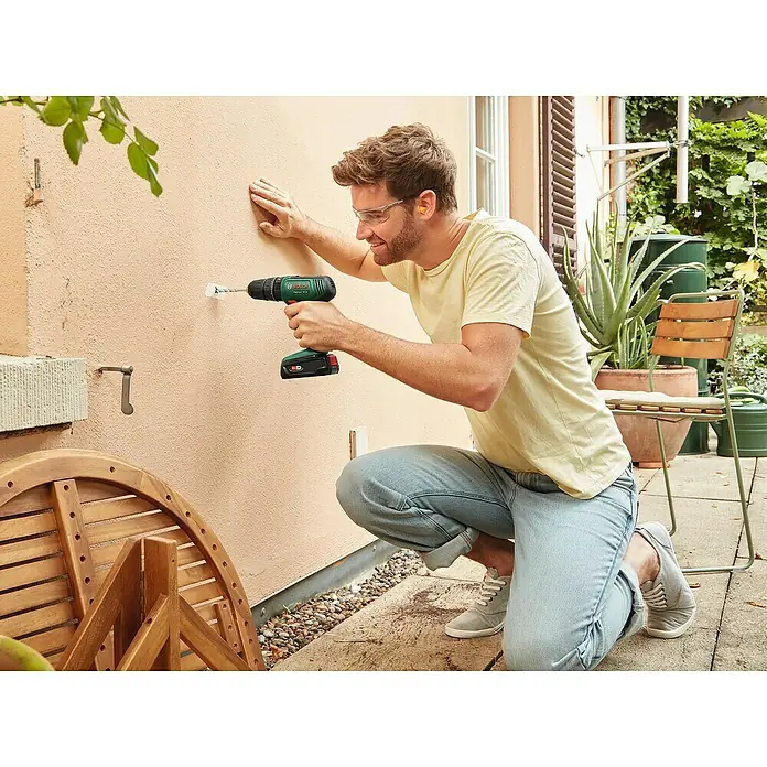 Bosch Power for All 18V Akku-Schlagbohrschrauber EasyImpact 18V-38 - 18 V, 1 Akku, 2 Ah, 38 Nm - Ein Mann bohrt mit einer grünen Bosch Akku-Bohrmaschine ein Loch in eine Wand, während er Sicherheitsbrille und Ohrstöpsel trägt.