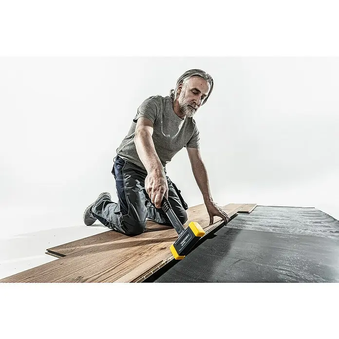 Halder Secural Schonhammer - Maße Kopf: 30 x 40 mm, Mittelhart - Mann verlegt mit einem SECURAL-Hammer Holzboden auf einer schwarzen Unterlage. Das Werkzeug ist gelb/schwarz und dient zur Verlegung.