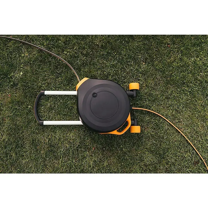Fiskars X-series Automatische Schlauchtrommel XL mit Rädern - Schlauchlänge: 25 m - Gartenschlauchaufroller auf Rasen. Der Schlauchaufroller ist schwarz mit orangefarbenen Akzenten und hat einen weißen Griff.
