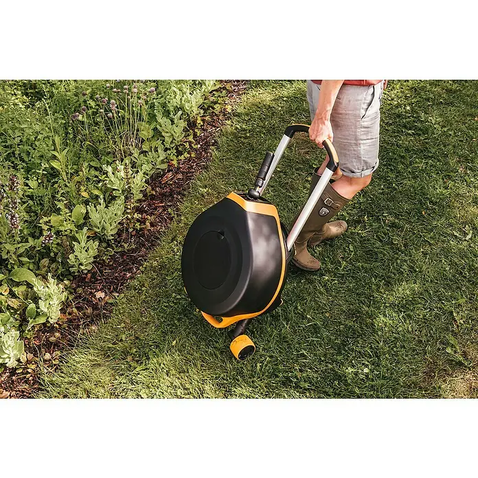 Fiskars X-series Automatische Schlauchtrommel XL mit Rädern - Schlauchlänge: 25 m - Eine Person schiebt einen schwarzen und gelben Gartenschlauchwagen auf einem grünen Rasen in einem Garten.