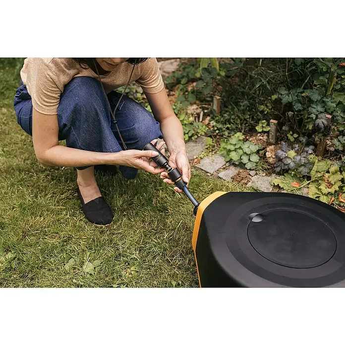 Fiskars X-series Automatische Schlauchtrommel XL - Schlauchlänge: 25 m - Eine Frau schliesst einen schwarzen und goldenen Gartenschlauch von Fiskars an, während sie auf einer Wiese kniet.