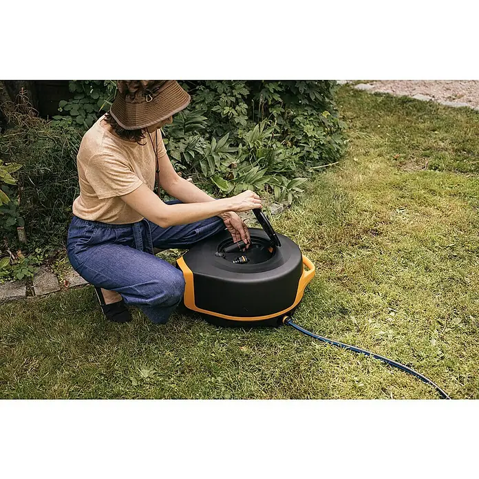 Fiskars X-series Automatische Schlauchtrommel XL - Schlauchlänge: 25 m - Eine Person hockt und benutzt eine schwarze Schlauchtrommel mit gelben Akzenten, die auf einer grünen Rasenfläche steht.