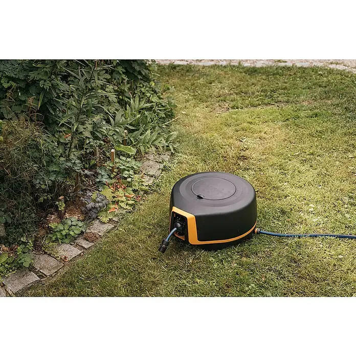 Fiskars X-series Automatische Schlauchtrommel XL - Schlauchlänge: 25 m - Ein schwarzer, runder Schlauchaufroller mit einem gelben Akzent sitzt auf einem Rasen neben einigen Pflanzen.