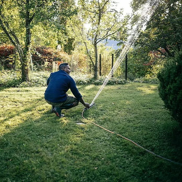 Fiskars X-series Ersatzteil-Set - Max. Regnerfläche: 350 m², Passend für: Größere Flächen - Mann bewässert Rasen mit Gartensprenger. Der Rasensprenger sprüht Wasser in einem üppigen Garten mit Bäumen und Sträuchern.