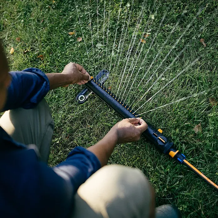 Fiskars X-series Ersatzteil-Set - Max. Regnerfläche: 350 m², Passend für: Größere Flächen - Ein Mann bedient einen oszillierenden Rasensprenger von Fiskars, der einen Rasen bewässert. Die Wasseraustrittsöffnungen sind gelb.