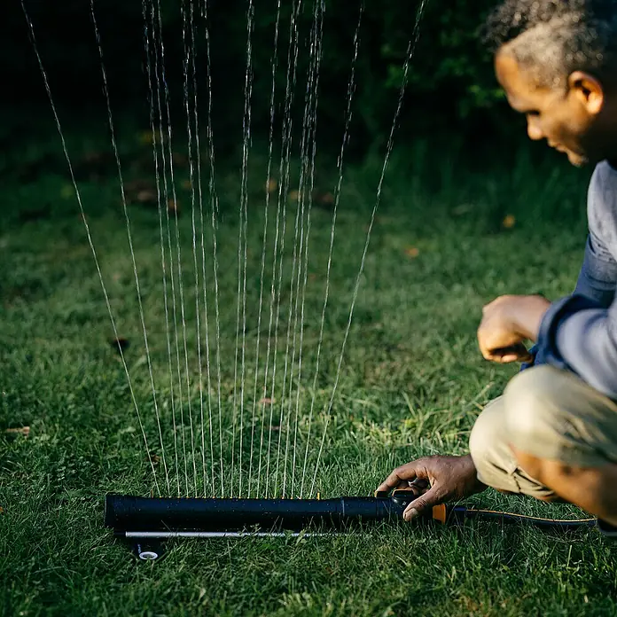 Fiskars X-series Ersatzteil-Set - Max. Regnerfläche: 350 m², Passend für: Größere Flächen - Ein Mann bedient einen Rasensprenger, der Wasserstrahlen auf eine grüne Rasenfläche sprüht. Der Sprenger ist schwarz und gelb.