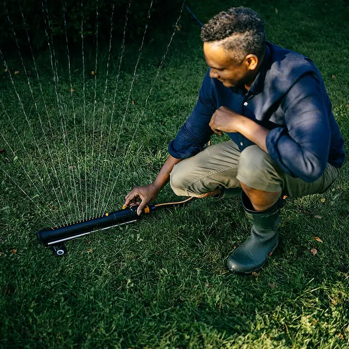 Fiskars X-series Ersatzteil-Set - Max. Regnerfläche: 300 m² - Mann stellt einen schwarzen Rasensprenger mit orangefarbenen Akzenten auf einer grünen Rasenfläche ein.