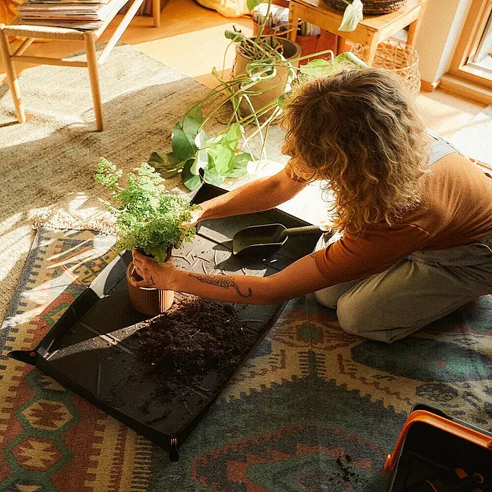 Fiskars Pflanzmatte - 96,8 x 57,8 cm - Eine Person kniet auf einem Teppich und topft eine Pflanze auf einer schwarzen Pflanzmatte um, mit Gartengeräten und Erde in der Nähe.