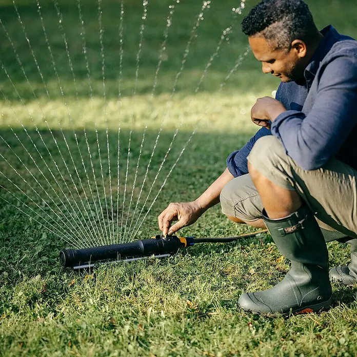 Fiskars Regner - Max. Regnerfläche: 300 m² - Ein Mann passt einen Rasensprenger im Garten an, Wasserstrahlen sprühen im Hintergrund. Fokus auf den Regner.