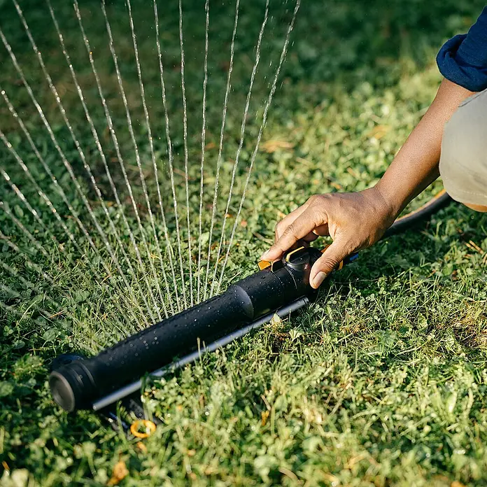 Fiskars Regner - Max. Regnerfläche: 300 m² - Der Handschuh reguliert den Wassersprenger auf einem grünen Rasen, wobei Wasserstrahlen in einem Bogen austreten. Der Wassersprenger ist schwarz und schlank.