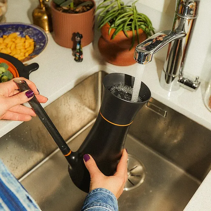 Fiskars Gießkanne - Schwarz, 1,2 l - Ein schwarzer Wasserfilterkrug mit einem orangefarbenen Band wird unter einem laufenden Wasserhahn in einer Spüle befüllt.