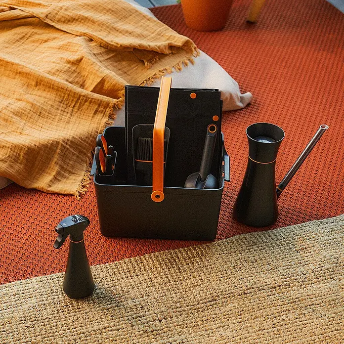 Fiskars Handsprüher - 0,3 l, Schwarz - Gartengeräte-Set mit schwarzer Gießkanne, Sprühflasche und Werkzeugkasten mit orangefarbenen Akzenten auf einem gemusterten Teppich.