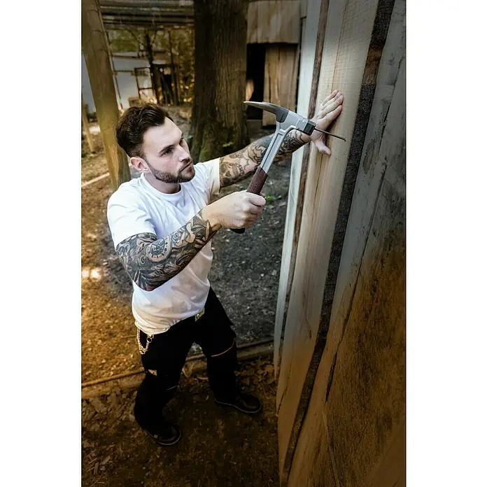 Picard Latthammer AluTec® - Gewicht Kopf: 750 g, Aluminium - Ein Zimmermann mit Tätowierungen an den Armen, der ein Nagel mit einem Hammer in ein Holzbrett schlägt.