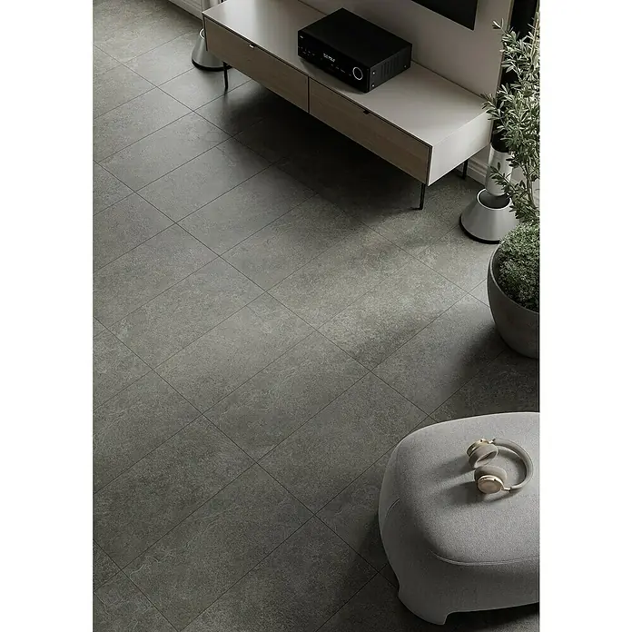 Feinsteinzeugfliese Ardezia - 29,8 x 59,8 cm, Graphite, Matt - Hochwinkelaufnahme eines minimalistischen Wohnzimmers mit grauen Fliesen, einem TV-Schrank, einer Zimmerpflanze und Kopfhörern auf einem Pouf.