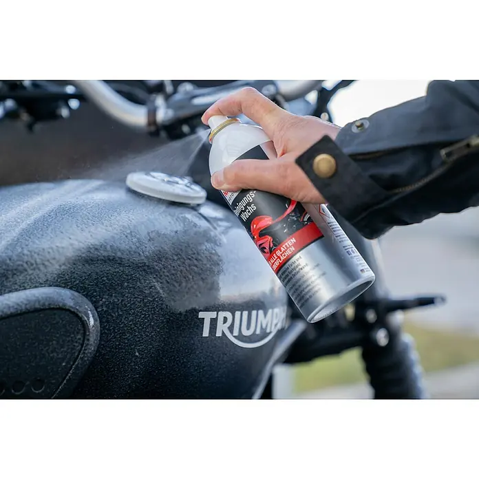 Nigrin Moto-Bike Sprühwachs - 400 ml - Eine Person sprüht Motorradreinigungswachs auf einen schwarzen Triumph-Tank, um ihn zu reinigen und zu schützen.
