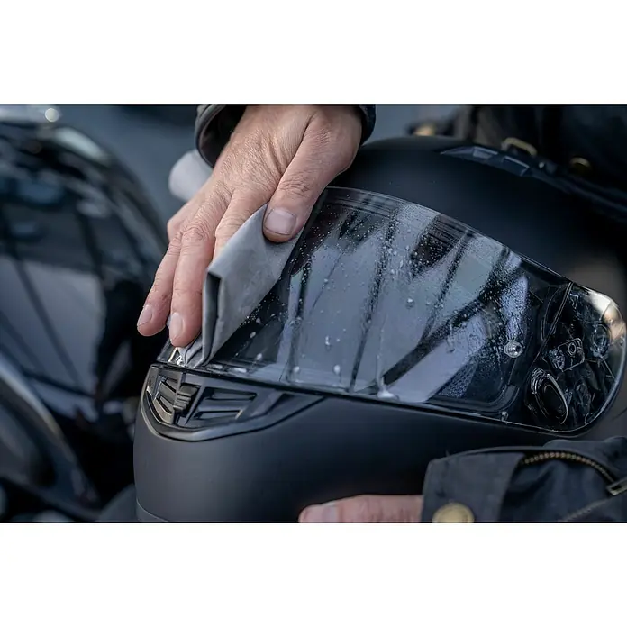 Nigrin Moto-Bike Mikrofaser-Reinigungstuch - 1 Stk. - Eine Hand wischt das Visier eines schwarzen Motorradhelms mit einem grauen Tuch ab, um die Feuchtigkeit zu entfernen.