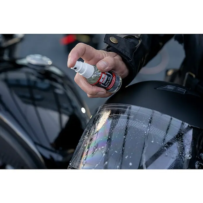 Nigrin Moto-Bike Reinigungsmittel - 50 ml, Sprühkopf - Sprühflasche Nigrin Helm- und Visierreiniger in der Anwendung auf einem Motorradhelmvisier, um klare Sicht und Sicherheit zu gewährleisten.