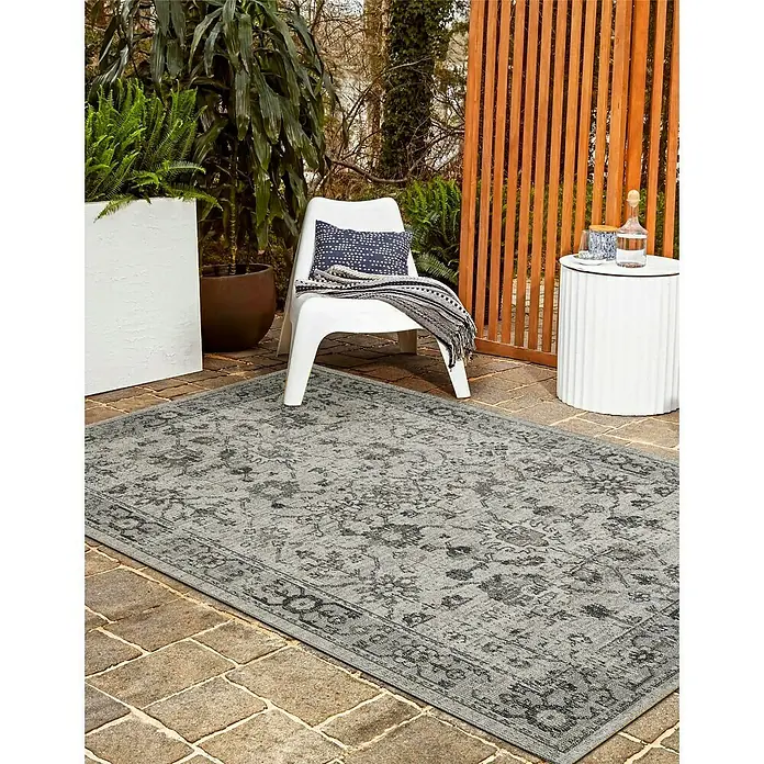 Outdoor-Teppich Austin - Hellgrau, 190 x 133 cm, 100 % Polypropylen - Ein grauer Outdoor-Teppich mit floralem Muster liegt auf einer Terrasse mit einem weißen Stuhl und einem Beistelltisch mit Getränken.