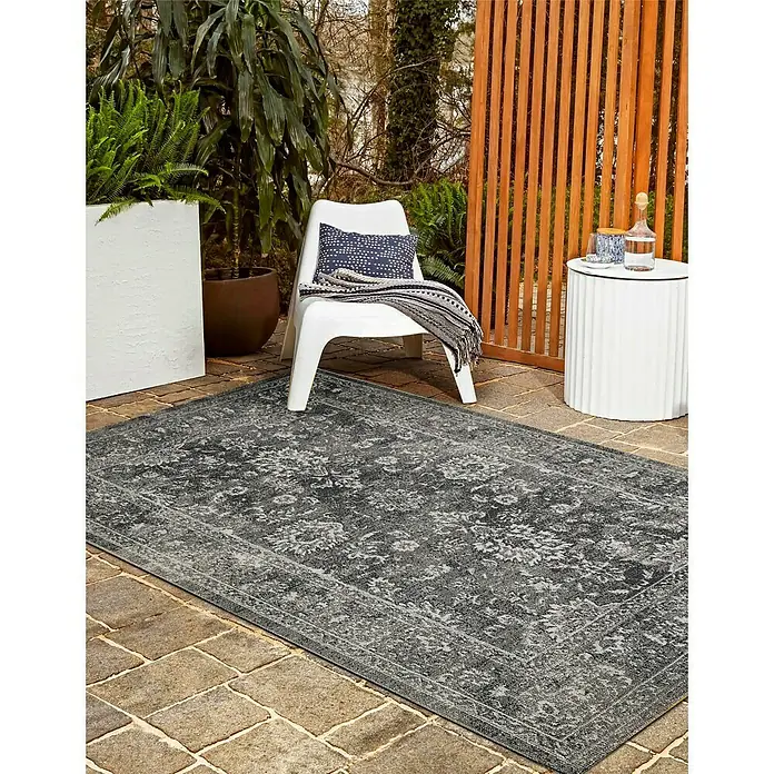 Outdoor-Teppich Austin - Grau, 190 x 133 cm, 100 % Polypropylen - Ein grauer Outdoor-Teppich mit floralem Muster liegt auf einer Terrasse neben einem weißen Stuhl und einem Beistelltisch.