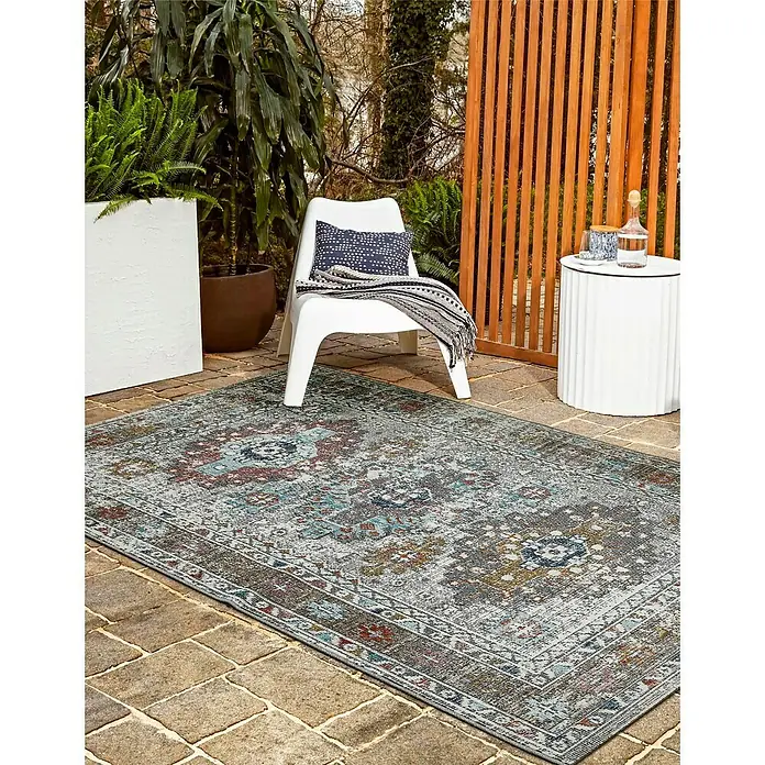 Outdoor-Teppich Austin - Multi, 190 x 133 cm, 100 % Polypropylen - Ein dekorativer, grauer Teppich mit einem traditionellen Muster liegt auf einer Terrasse, umgeben von Pflanzen und Gartenmöbeln.