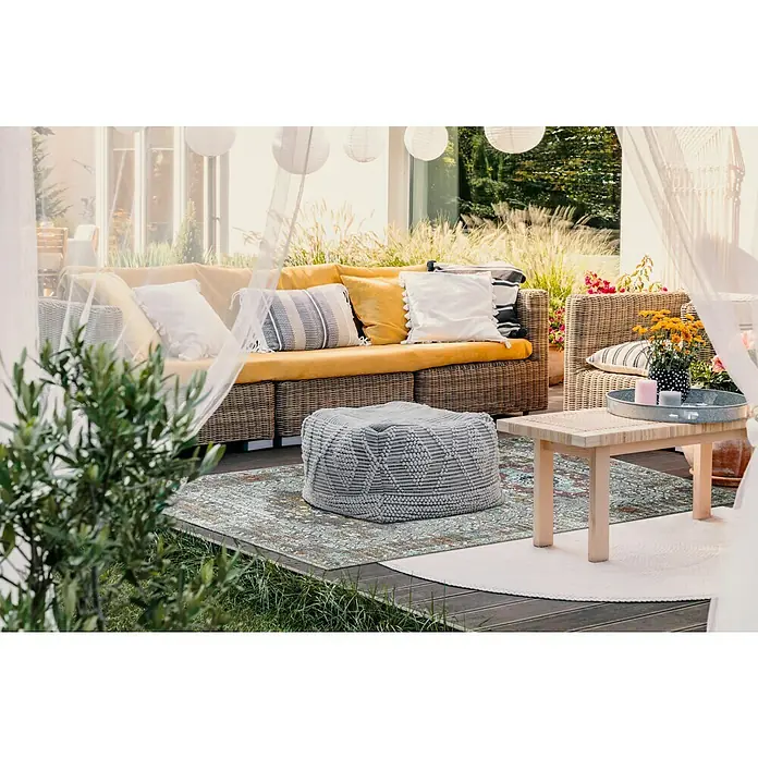 Outdoor-Teppich Austin - Multi, 190 x 133 cm, 100 % Polypropylen - Ein stilvolles Außenwohnzimmer mit einem Outdoor-Teppich mit kunstvollen Mustern unter einem Ottomane und einem Holztisch.