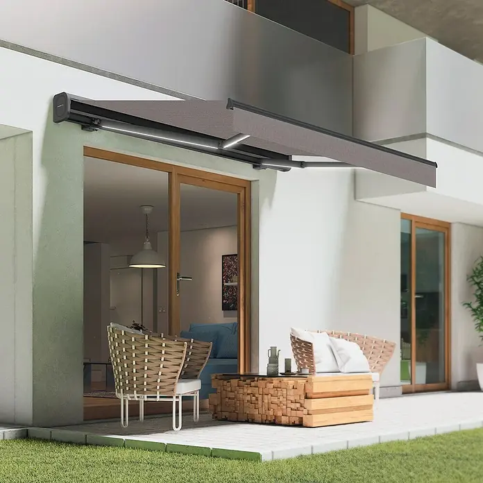 SmartSun Toldo con motor Dark Vivant - Gris piedra, Ancho: 4,75 m, Caída: 3 m - Un moderno toldo de patio con iluminación LED proporciona sombra sobre una zona de estar al aire libre, creando un ambiente agradable.