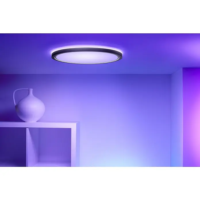 Philips Wiz Smart-LED-Deckenleuchte rund Tunable White & Color SuperSlim - 32 W, Ø x H: 55 x 4,8 cm, Schwarz, RGBW - Deckenleuchte in einem Raum mit lila Beleuchtung, neben einem Regal mit einer weißen Vase. Die Lampe ist rund und leuchtet hell.