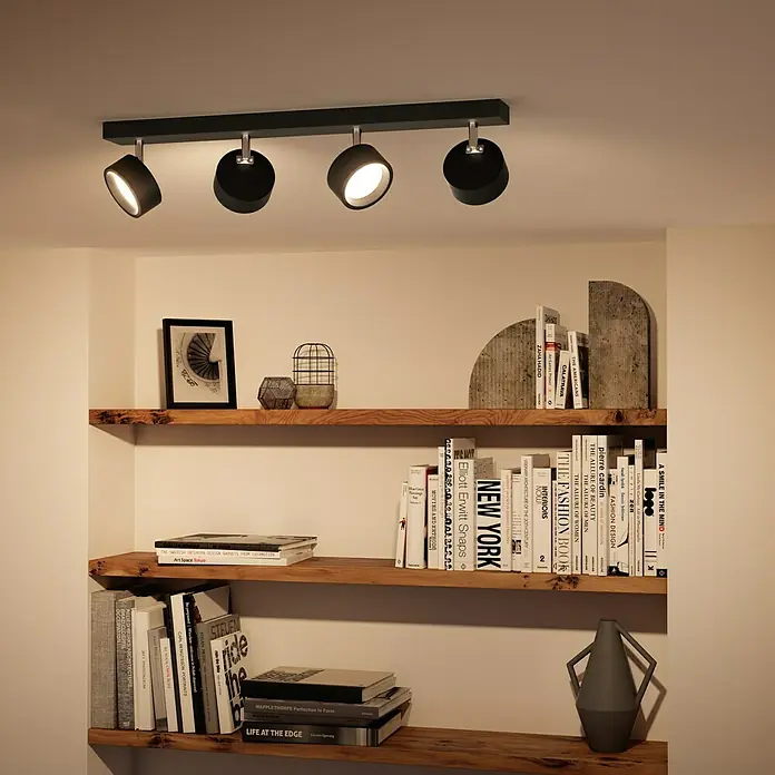 Philips Regleta LED Bracia - 22 W, L x An x Al: 60 x 8 x 12,5 cm, Negro, Blanco cálido, 4 ud. - Una lámpara de techo negra moderna con cuatro focos ilumina una estantería llena de libros y objetos decorativos.