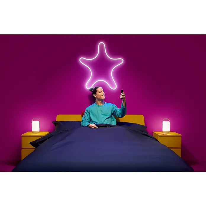 Philips Wiz Smart-LED-Band - Länge: 5 m, RGB, 24 W, 240 lm, 220 V - 240 V - Mann liegt im Bett mit blauem Bettzeug und gelbem Nachttisch und benutzt ein Mobiltelefon unter einer leuchtenden Sternenlampe.