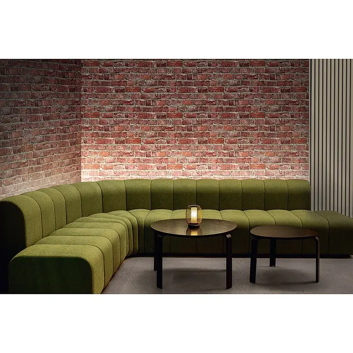 Marburg GZSZ Vliestapete Steinwand - Ziegelrot, Steinoptik, 10,05 x 0,53 m - Einladende Sitzecke mit einem modularen grünen Sofa, dunklen Holztischen und einem warm leuchtenden Windlicht vor einer Ziegelwand.