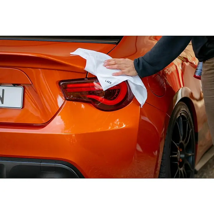 Nigrin Performance Mikrofaser-Set - 3 -tlg. - Nahaufnahme eines orangefarbenen Autos, das mit einem weißen "LACK"-Tuch poliert wird, wodurch die glänzende Lackierung hervorgehoben wird.