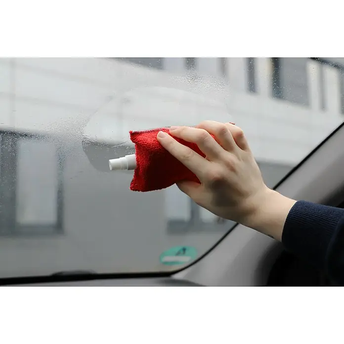 Nigrin Antibeschlagschwamm - Ausstattung: Sprühkopf - Das Bild zeigt eine Hand, die mit einem roten Tuch und einer Sprühflasche eine Autoscheibe putzt, um sie von Beschlag zu befreien.