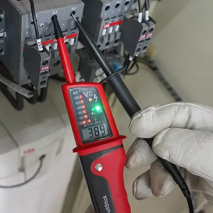 Multímetro digital UT15C - 12 V - 690 V - Probador eléctrico rojo y negro sostenido por una mano enguantada, mostrando 389 voltios en componentes eléctricos grises.