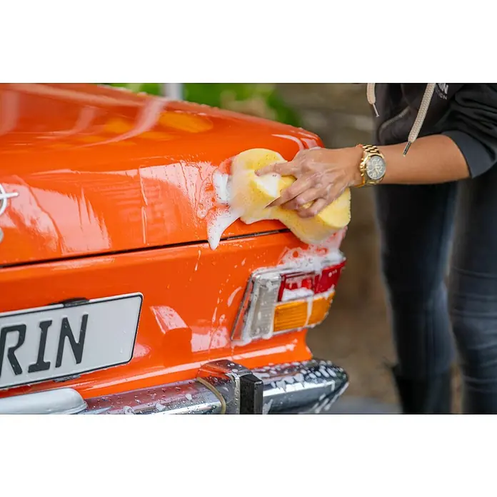 Nigrin Autoschwamm Super Soft - 19 x 14 mm - Nahaufnahme einer Person, die ein orangefarbenes Auto mit einem gelben Schwamm und Seife wäscht, um es sauber und glänzend zu halten.