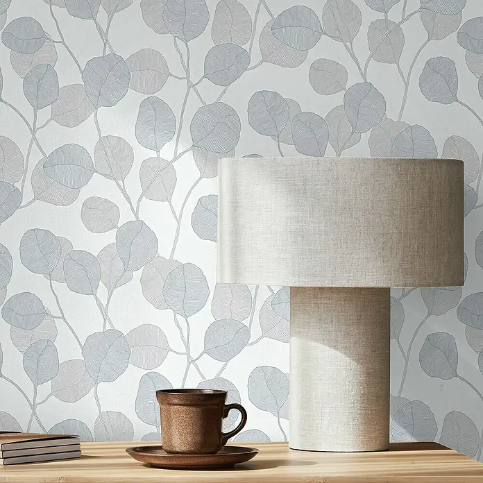 Marburg GZSZ Vliestapete Blätterranke - Weiß/Graublau, Floral, 10,05 x 0,53 m - Eine moderne Tischleuchte mit beigem Lampenschirm auf einem Holztisch mit Tasse und Notizbuch, vor einer Wand mit Blättermuster.
