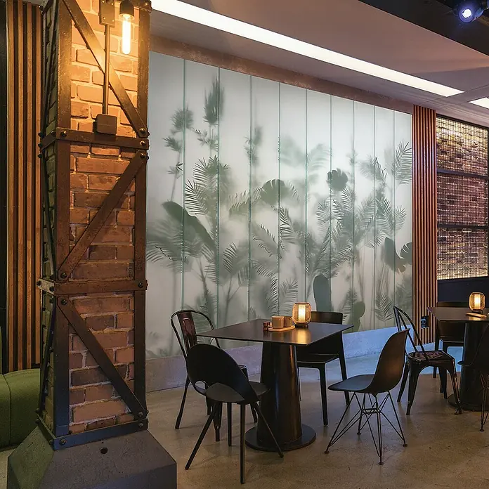 Marburg GZSZ Fototapete Tropische Pflanzen - B x H: 1,59 x 2,7 m - Elegantes Café-Interieur mit dunklen Tischen und Stühlen, akzentuiert durch eine dekorative Backsteinwand und indirekte Beleuchtung.