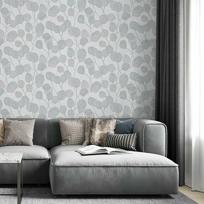 Marburg GZSZ Vliestapete Blätterranke - Weiß/Graublau, Floral, 10,05 x 0,53 m - Elegantes Interieur mit einer grauen modularen Couch, dekoriert mit Kissen, vor einer Wand mit einem dezenten Blattmuster.