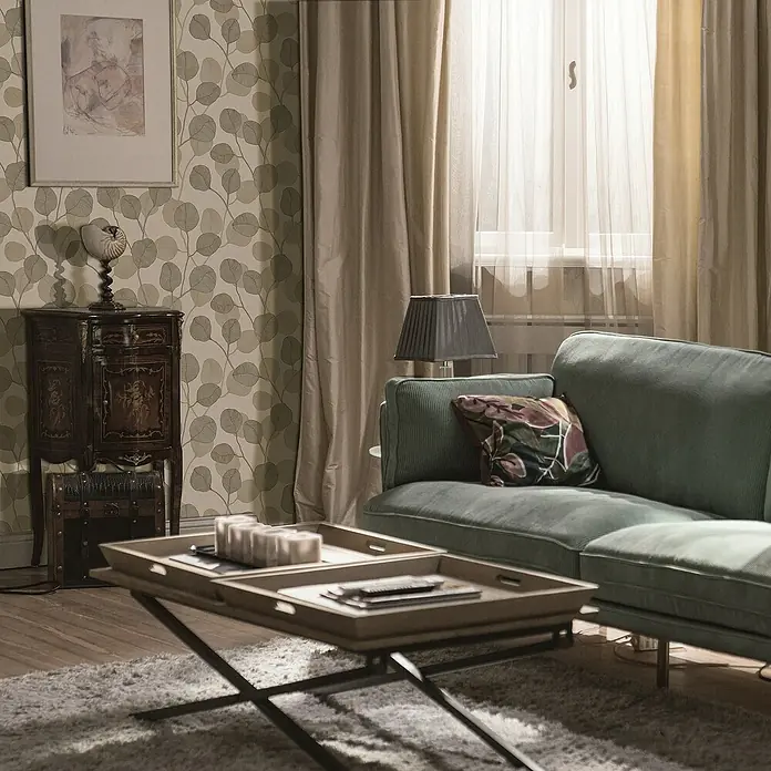 Marburg GZSZ Vliestapete Blätterranke - Beige/Braun, Floral, 10,05 x 0,53 m - Stilvolles Wohnzimmer-Interieur mit einem grünen Cord-Sofa, einem Couchtisch mit Tabletts und einer eleganten Kommode vor einer gemusterten Tapete.