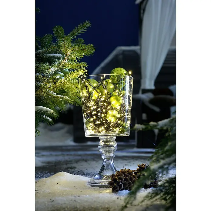 Tween Light LED-Lichterkette - Außen, Lichtfarbe: Warmweiß, Anzahl Leuchtmittel: 200 Stk., Netzbetrieben, 35 m - Eine stilvolle Glasvase, gefüllt mit grünen Kugeln und warmen Lichtern, steht im Schnee, umgeben von Tannenzweigen.