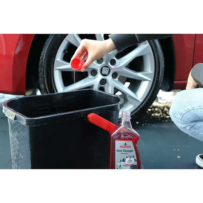 Nigrin Auto-Shampoo Konzentrat - 1 l, Orangenduft - Eine Person schüttet Nigrin Auto-Shampoo-Konzentrat in einen Eimer vor einem roten Auto, um es zu waschen.