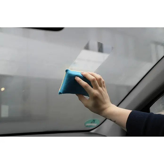 Nigrin Antibeschlagschwamm - 2-seitig - Person, die mit einem blauen und gelben Schwamm eine Autoscheibe reinigt, um klare Sicht zu gewährleisten. Perfekt für die Autopflege.
