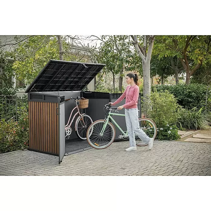 Keter Truhe Signature Venetian Grande Store - L x B x H: 109,3 x 190,5 x 132 cm, Braun - Dunkelgrauer Outdoor-Stauraum mit Holzoptik-Paneelen, offen, eine Frau verstaut ein Fahrrad, ein weiteres ist bereits drinnen, im Garten.
