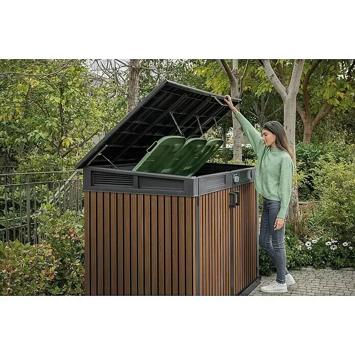 Keter Truhe Signature Venetian Grande Store - L x B x H: 109,3 x 190,5 x 132 cm, Braun - Outdoor-Mülltonnenbox in Holzoptik mit dunkelgrauem Rahmen, geöffnet mit zwei grünen Behältern, von einer Frau bedient.