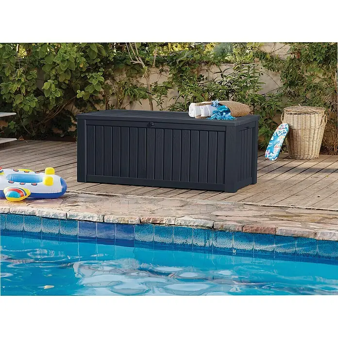 Keter Gartenbox Rockwood - 570 l, Kunststoff - Eine dunkelblaue Gartenbox aus Kunststoff mit vertikalen Brettern steht auf einer Holzterrasse neben einem Pool. Praktische Aufbewahrung für den Außenbereich.