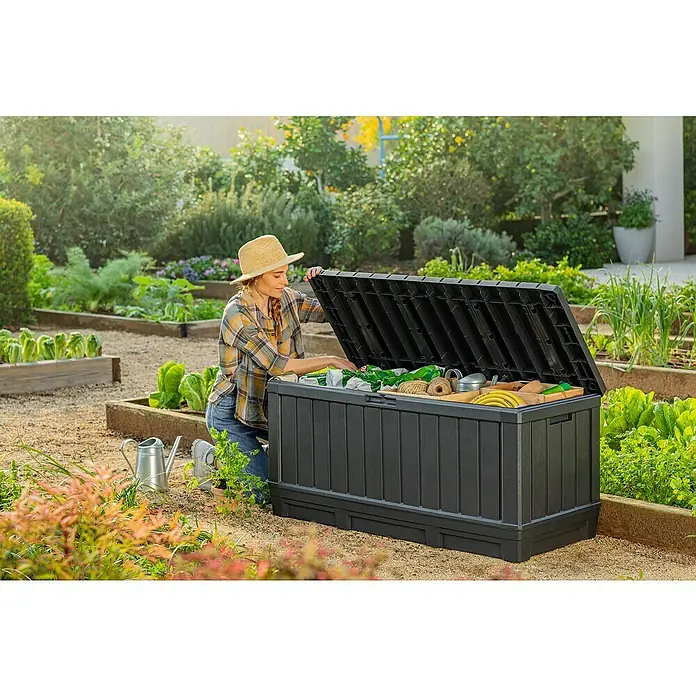 Keter Garten-Aufbewahrungsbox Kentwood - L x B x H: 128 x 53,6 x 59 cm, Kunststoff - Eine Frau kniet neben einer schwarzen Gartenbox, die mit Gartengeräten gefüllt ist, in einem üppigen Garten.