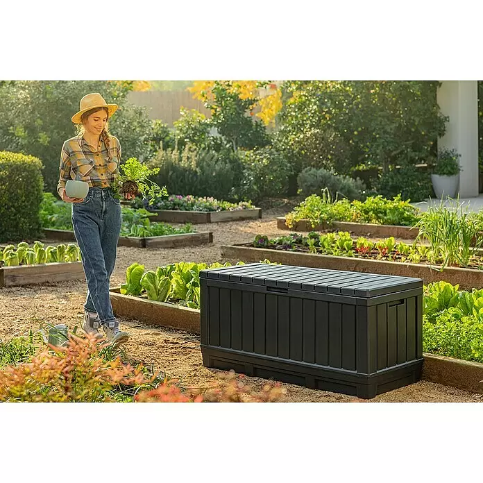 Keter Arcón para cojines de exterior Kentwood - L x An x Al: 128 x 53,6 x 59 cm, Plástico, Gris - Caja de almacenamiento exterior gris oscuro con tapa de listones, situada en un jardín entre bancales elevados.