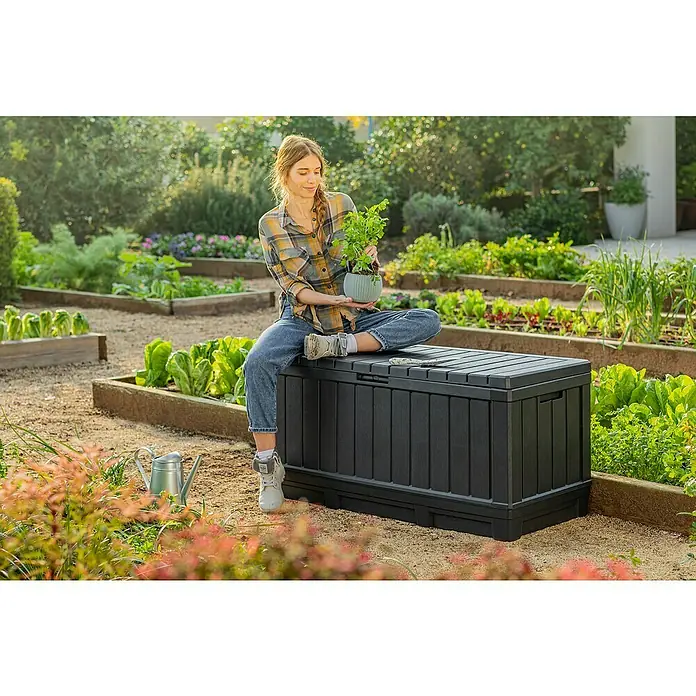 Keter Arcón para cojines de exterior Kentwood - L x An x Al: 128 x 53,6 x 59 cm, Plástico, Gris - Una joven sentada en una caja de almacenamiento de jardín negra, sosteniendo una planta en maceta, rodeada de un jardín exuberante.