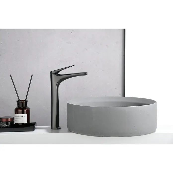 Imex Delos Grifo de lavabo alto - Black gun metal - Primer plano de un lavabo gris con grifo, difusor de aroma y producto para el cuidado de la piel sobre un tocador blanco contra una pared gris.