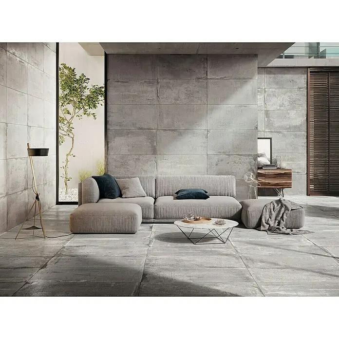 Feinsteinzeugfliese Bond Ash - 60 x 120 cm, Grau, Matt - Ein modernes Wohnzimmer mit grauem Sofa, Couchtisch und Stehlampe. Die grauen Fliesen und die Betonwand erzeugen ein elegantes Ambiente.