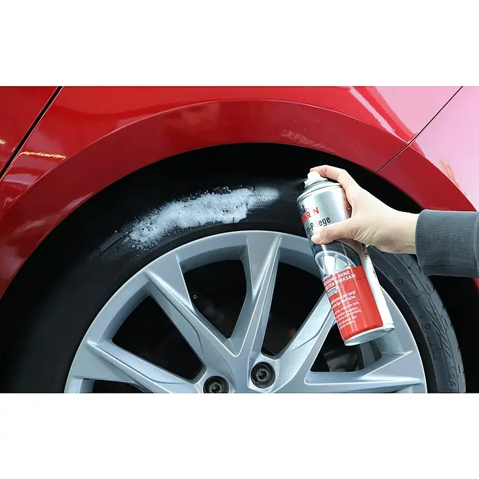 Nigrin Reifenpflege - 500 ml - Ein rotes Auto mit einer Person, die Reifenschaum auf einen Reifen sprüht, um ihn zu reinigen. Der Reifenschaum sorgt für eine saubere Oberfläche.